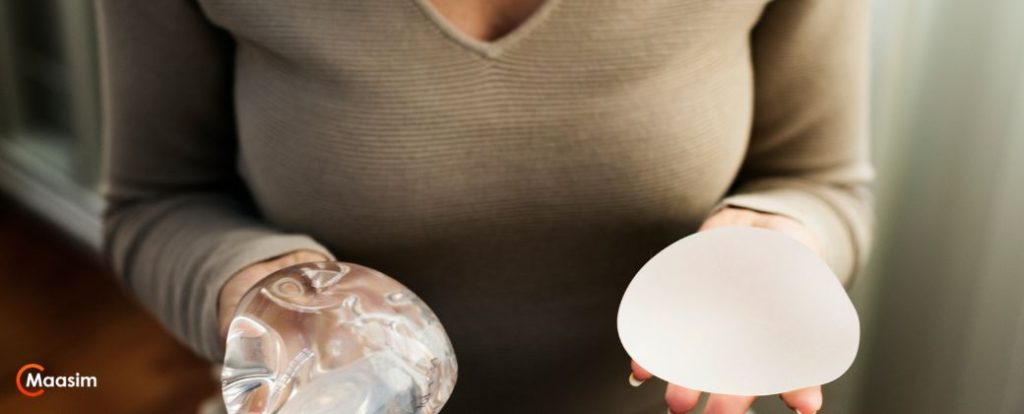 שתלי סיליקון – מה באמת חשוב לדעת לפני הניתוח? - תמונת פלג גוף עליון של אישה הלובשת סריג אפרפר ומחזיקה שתל בכל יד ובצד שמאל למטה לוגו אתר MAASIM